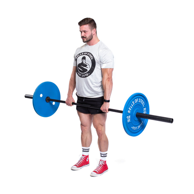 A man wearing a gray T-shirt and black shorts is holding a barbell equipped with Bells of Steel Calibrated Powerlifting Plates - KG. He stands on a white background, sporting red high-top sneakers with white accents. The blue color-coded plates are designed to meet IPF specs for precise training sessions.