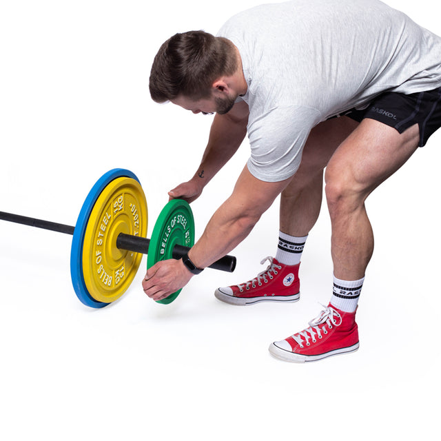 A man in a gray shirt and red sneakers is adjusting weights on a barbell. The Bells of Steel Calibrated Powerlifting Plates - KG, which are color-coded in yellow, blue, and green to meet IPF specifications, are featured. He is standing on a white surface.