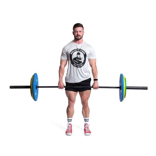 A person stands holding a barbell with vibrant, colour-coded Bells of Steel Calibrated Powerlifting Plates - KG that meet IPF specs. They're dressed in a gray T-shirt, black shorts, white socks, and red sneakers against a plain white background.