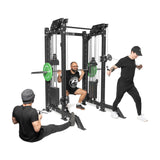 Three people utilize THE KOLOSSOS - Smith Functional Trainer by Bells of Steel in their home gym: one squats with the barbell on the Smith Machine, another uses the pulley system while standing, and a third sits with a phone. All are dressed in athletic wear.