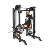 A person with a beard works out on THE KOLOSSOS - Smith Functional Trainer by Bells of Steel in their home gym. Dressed in a logoed black t-shirt, black shorts, knee sleeves, and glasses, they use the large machine with multiple cables and weight stacks for an effective workout.