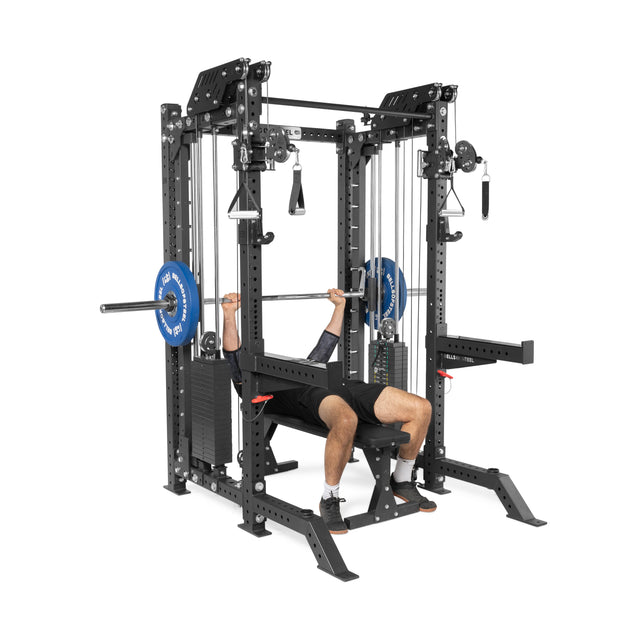 In their home gym, a person uses THE KOLOSSOS - Smith Functional Trainer by Bells of Steel for bench pressing. They lie on a bench, gripping a barbell overhead. The trainer's pulleys and weight stack highlight the versatility of this fitness equipment.