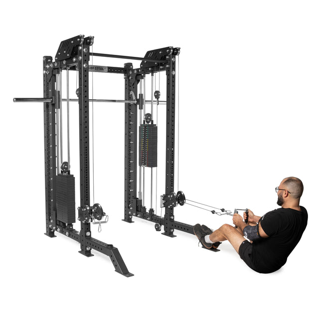 A man in a black shirt and shorts uses THE KOLOSSOS - Smith Functional Trainer by Bells of Steel to perform a seated cable row exercise. This versatile equipment, featuring pulleys and weight stacks, is ideal for home gyms and fitness centers, providing an effective workout with a cable-attached handle.