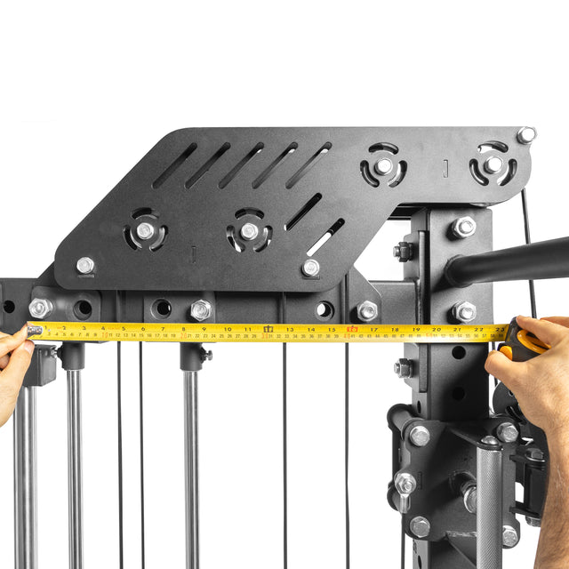 Someone measures a black metal exercise machine, likely part of a home gym, with a yellow tape measure. The image highlights the upper section of Bells of Steel's THE KOLOSSOS - Smith Functional Trainer, featuring various metal plates, pulleys, and bolts.