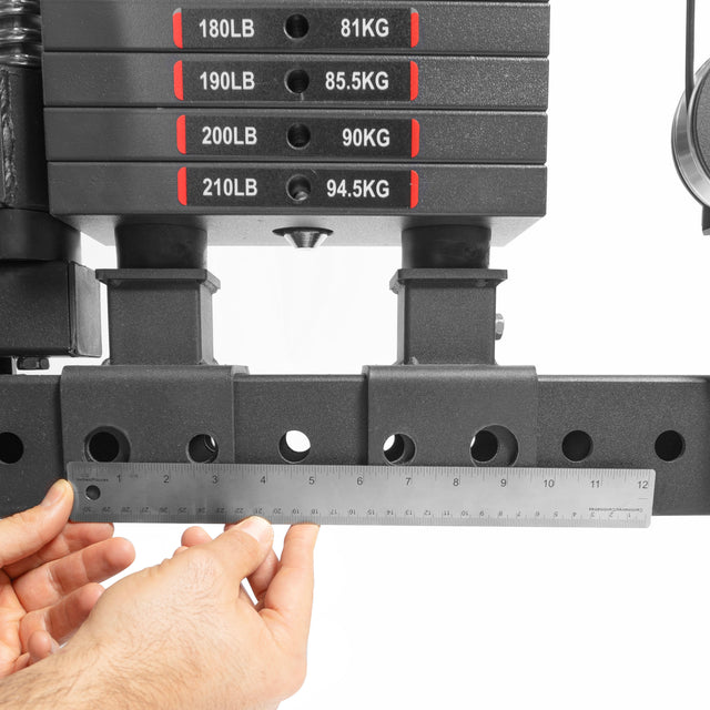 Close-up of hands holding a ruler under stacked weight plates from THE KOLOSSOS - Smith Functional Trainer by Bells of Steel. Weights range from 180 to 210 pounds with kilograms shown, measuring the gap beneath.