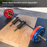 The Bells of Steel Safety Squat Bar - The SS4, with padded shoulder rests and colorful weight plates, sits on an outdoor wooden lifting platform. Text reads: “Voted ‘Best Versatile Safety Squat Bar’ – King Of The Gym.”.