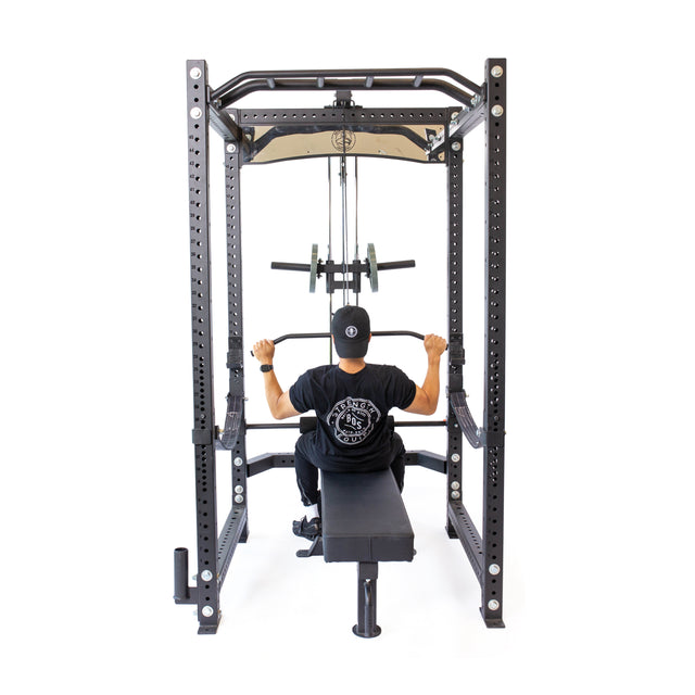 A person in a black T-shirt and cap is using the Bells of Steel Lat Pulldown & Low Row Rack Attachment - Hydra & Manticore in the gym, pulling down the bar with both hands on this versatile black metal machine featuring various cable exercise attachments.