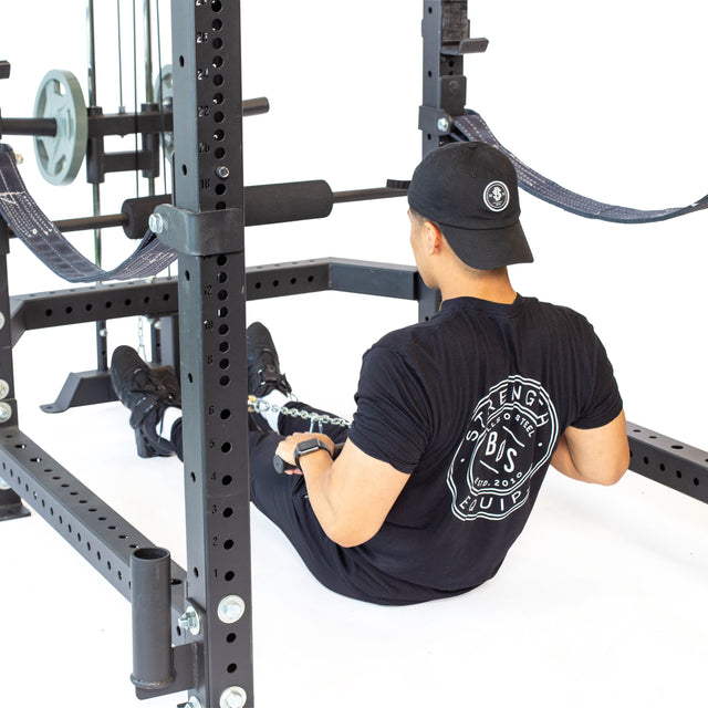 A person in a black cap and t-shirt with a circular logo performs cable exercises on the gym floor, surrounded by Olympic weight plates and a Bells of Steel Lat Pulldown & Low Row Rack Attachment - Hydra & Manticore, enhancing the workout atmosphere.