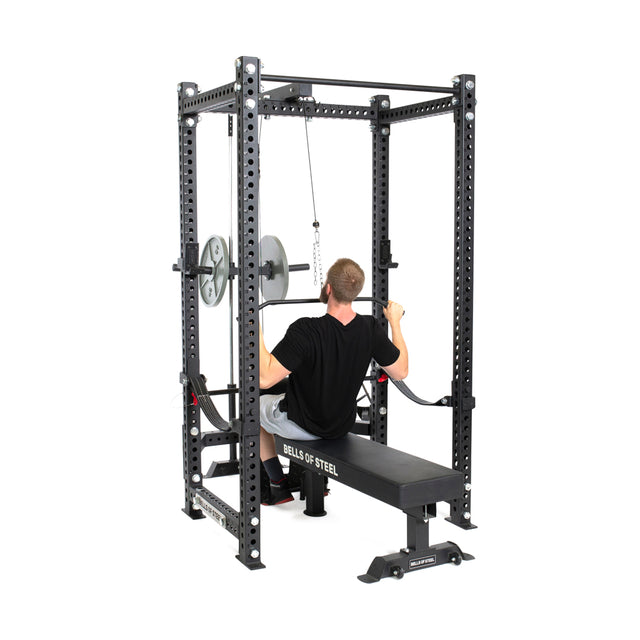 A person performs a lat pulldown on a bench within a steel power rack, utilizing the Lat Pulldown & Low Row Rack Attachment from Bells of Steel.