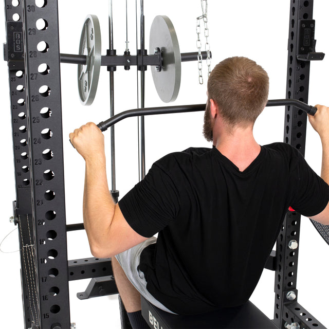 A short-haired man with a beard is on a gym bench doing lat pull-downs with the Bells of Steel Lat Pulldown & Low Row Rack Attachment - Hydra & Manticore, using Olympic weight plates and chains. He's wearing a black T-shirt and light shorts.