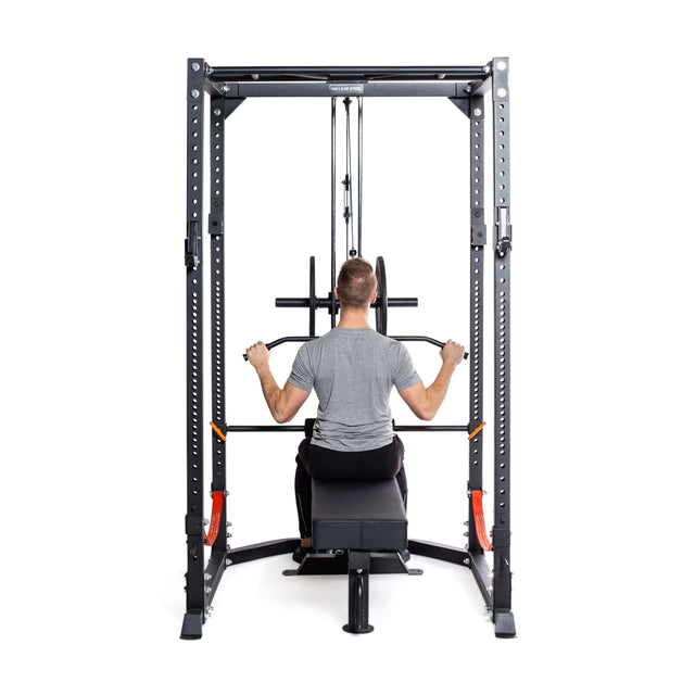 A man is using the Bells of Steel Rack Lat Pulldown/Row Attachment for a lat pulldown workout. He's in a gray T-shirt and black shorts, seated on the sleek black-framed machine with red and silver accents, featuring an advanced cable pulley system.