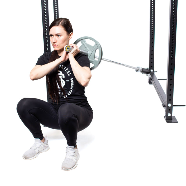 In a gym, a long-haired individual in a black workout outfit and white sneakers performs a front squat with a barbell. They utilize the Bells of Steel Landmine Rack Attachment to demonstrate intense strength workouts against the plain white background.