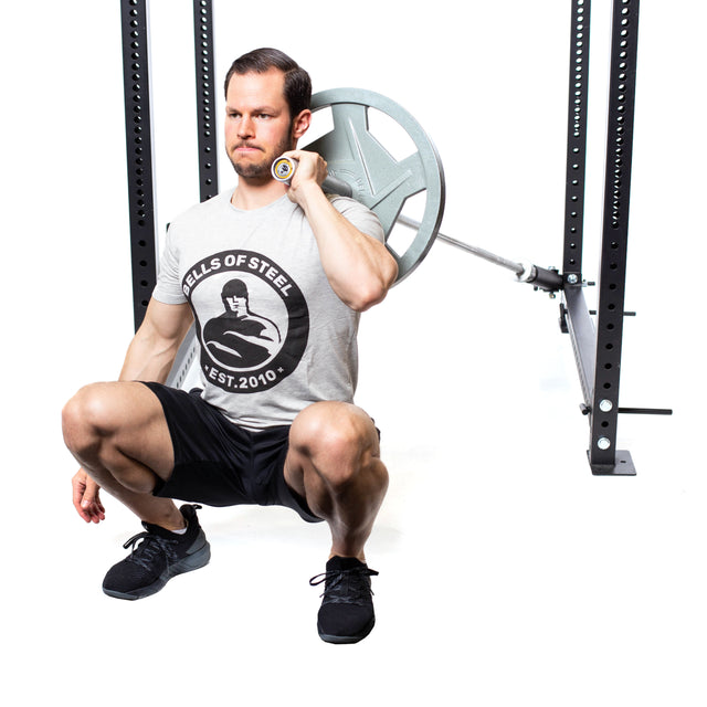 A man performs a squat with a barbell on his shoulders, wearing a gray T-shirt, black shorts, and black sneakers. Utilizing the Bells of Steel Landmine Rack Attachment for stability and rotational core work against a minimalist white background.