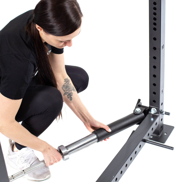 Dressed in black with white sneakers and a rose tattoo on their right forearm, a person squats to attach a Bells of Steel Landmine Rack Attachment to a squat rack with UHMW protection. The equipment's numbered holes make it ideal for strength workouts.