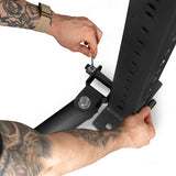 A tattooed individual uses a wrench to secure bolts on a large black Landmine Rack Attachment by Bells of Steel. The adjustable frame, designed with holes for height modifications, is ideal for intense workouts and protecting Olympic barbells.