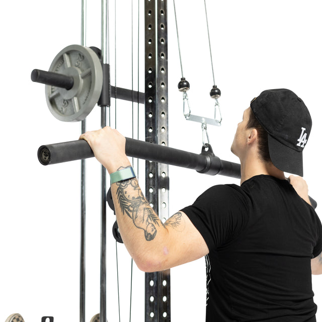 A person with tattoos on their left arm, wearing a black t-shirt and cap, performs a lat pulldown using the Bells of Steel Fat Bar - Lat Pulldown Bar, gripping it with both hands. Gym weights are loaded on the machine.