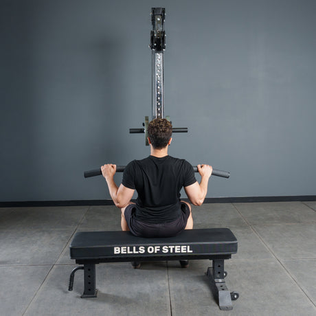 A person sits on a Bells of Steel bench, gripping the Fat Bar - Lat Pulldown Bar attached to a lat pulldown machine, ready to boost grip strength in a gym with gray walls and flooring.