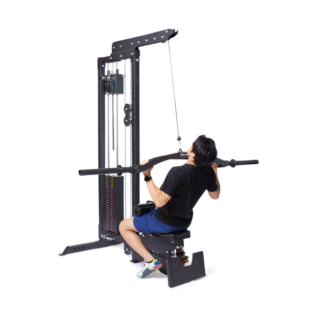 A person in a black shirt and blue shorts uses the Bells of Steel Lat Pulldown Low Row Machine in their home gym. Seated on the bench, they grip the bar with both hands, pulling it down to their chest with focused precision.