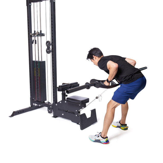 In a gym with a white background, someone in athletic wear uses the Bells of Steel Lat Pulldown Low Row Machine, performing a rowing exercise by pulling the bar while standing bent over.