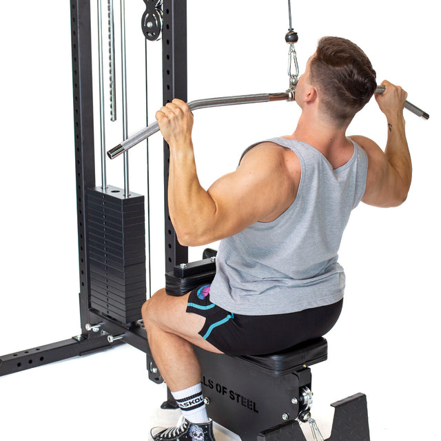 A person in a gray tank top and black shorts uses the Bells of Steel Lat Pulldown Low Row Machine at the gym, pulling down the bar with visible knee supports, testing their strength with each adjustment to the weight stack.