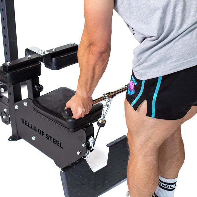 A person in a gray shirt and black shorts uses a Bells of Steel Lat Pulldown Low Row Machine, gripping the handle attached to cables. The focus is on their arm and leg muscles while working out in their home gym. The machine is black with visible weights and padding.