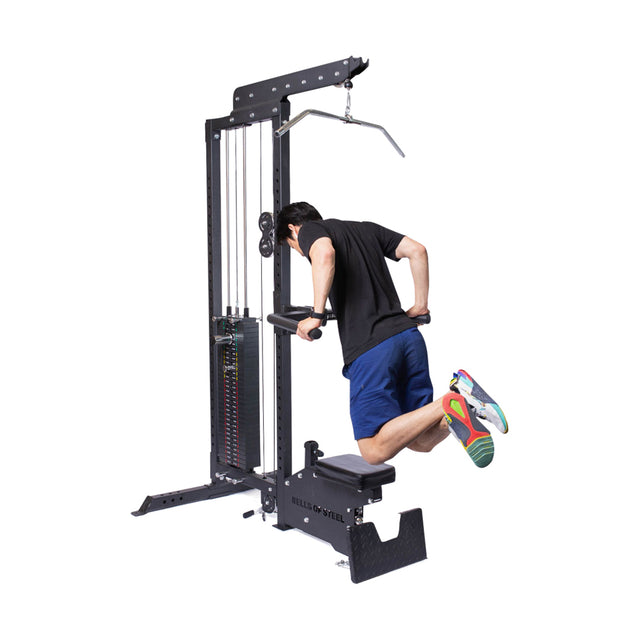 In a home gym, someone in a black shirt and blue shorts uses the Bells of Steel Lat Pulldown Low Row Machine for assisted dips. Equipped with weights and pulleys, they lean forward on the handles while sporting colorful sneakers for style.