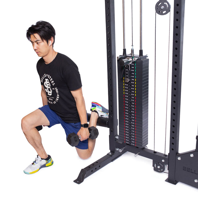 Using the Bells of Steel Lat Pulldown Low Row Machine for balance, a person performs a split squat in their home gym. They're dressed in a black t-shirt, blue shorts, and colorful sneakers while resting one foot on the machine's base for stability.