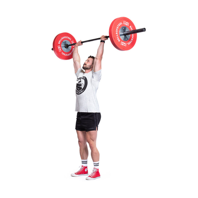 A person clad in a gray shirt and black shorts hoists a barbell overhead, equipped with Bells of Steel LB Competition Bumper Plates. They stand against a white backdrop, demonstrating their strength while sporting red shoes and white socks.