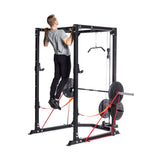 A man in a gray t-shirt and black pants does a pull-up on the Bells of Steel Light Commercial Power Rack (2.3" x 2.3", ⅝" holes) with resistance bands, safety straps, and weight plates attached, set against a white background.
