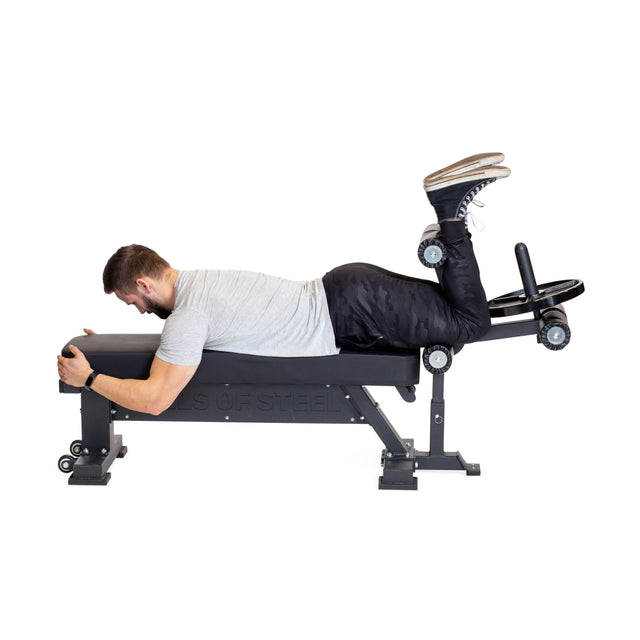 A person in a gray shirt and black pants is lying face down on the buzzsaw bench, performing a leg curl exercise with precision. Their knees are bent, and their feet are secured under padded rollers, highlighting the use of Bells of Steel's Bench Attachments for enhanced stability.