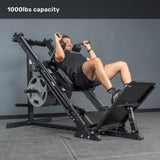 A man smiles while using the Bells of Steel Leg Press Hack Squat Machine, loaded with weight plates in a gym. Text at the top reads "1000lbs capacity." The machine has a sleek black design and is ideal for any home gym. Plain gray background.