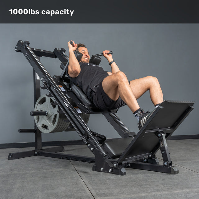 A man smiles while using the Bells of Steel Leg Press Hack Squat Machine, loaded with weight plates in a gym. Text at the top reads "1000lbs capacity." The machine has a sleek black design and is ideal for any home gym. Plain gray background.