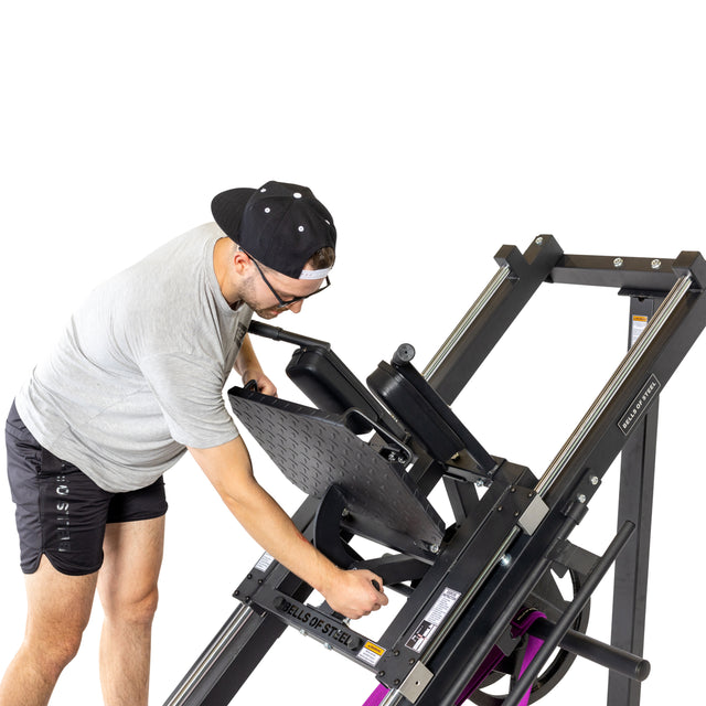 A person in a gray shirt, black shorts, and backward cap adjusts the sled capacity of the Bells of Steel Leg Press Hack Squat Machine, interacting with its seat and handles against a plain white background.