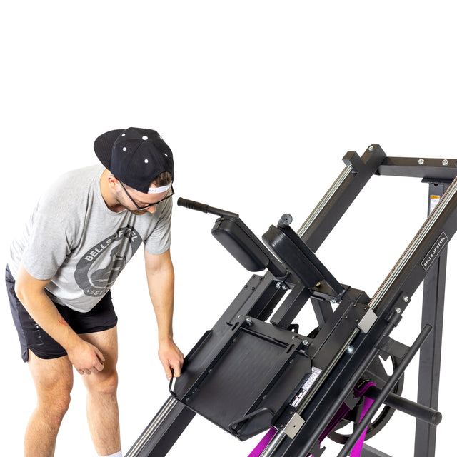 A person in athletic wear adjusts the sled capacity on a Bells of Steel Leg Press Hack Squat Machine at the gym, set against a plain white background highlighting both the equipment and their focused activity.