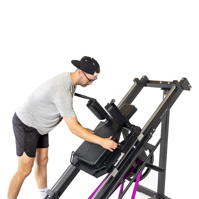 A person in a gray shirt, black shorts, and cap is adjusting a Bells of Steel Leg Press Hack Squat Machine with purple resistance bands. The setup subtly mimics the hack squat against a plain white background.