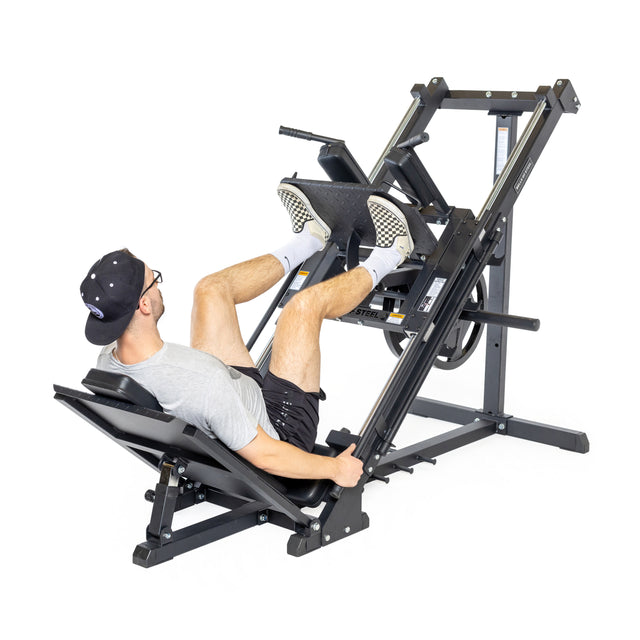 At the gym, someone in a gray t-shirt, black shorts, cap, and glasses is using the Bells of Steel Leg Press Hack Squat Machine. Seated with focus and determination, they push against the platform with precision.