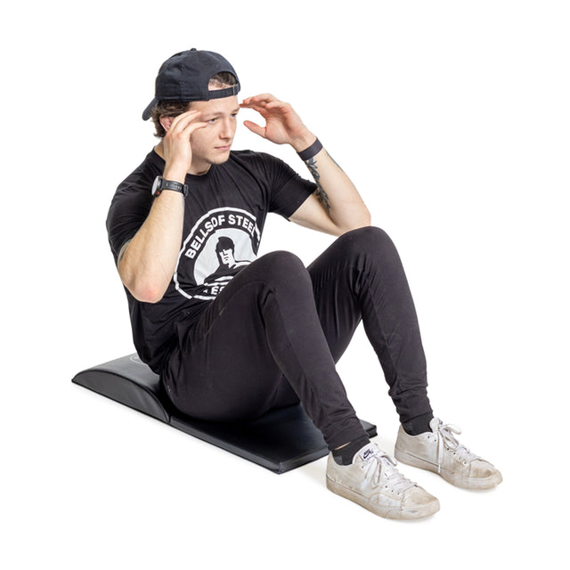 Wearing a black outfit and cap, a person performs a sit-up on the Bells of Steel Luxury Sit Up Mat, maintaining perfect lumbar curvature. Their hands are positioned by their head with bent elbows, while their white sneakers provide a crisp contrast to the scene.