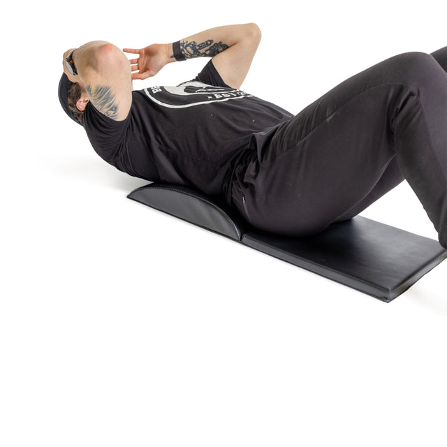 A person performs ab exercises on the Bells of Steel Luxury Sit Up Mat. They're lying back with knees bent, hands behind their head, wearing a black T-shirt and pants. Their tattooed right arm is visible as the white background highlights their form and lumbar curvature.