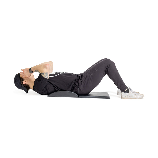 A person in a black outfit and cap performs an abdominal crunch on the Bells of Steel Luxury Sit Up Mat, designed to support lumbar curvature. Laying on this portable mat with knees bent and hands behind the head, they exercise effortlessly against a white background.