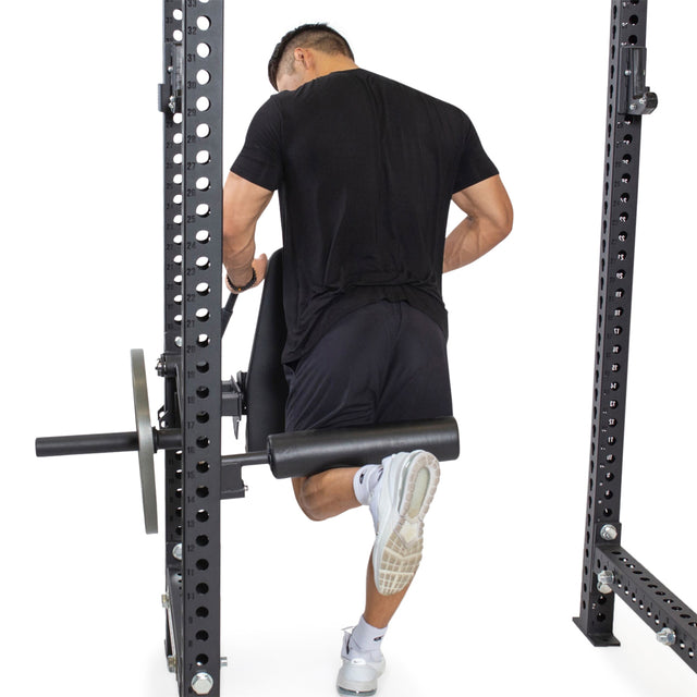 A person in a black shirt and shorts uses the Bells of Steel Leg Curl / Leg Extension Rack Attachment for a squat exercise. They're facing away with one leg on a padded bar and weights on each side against a plain white background.