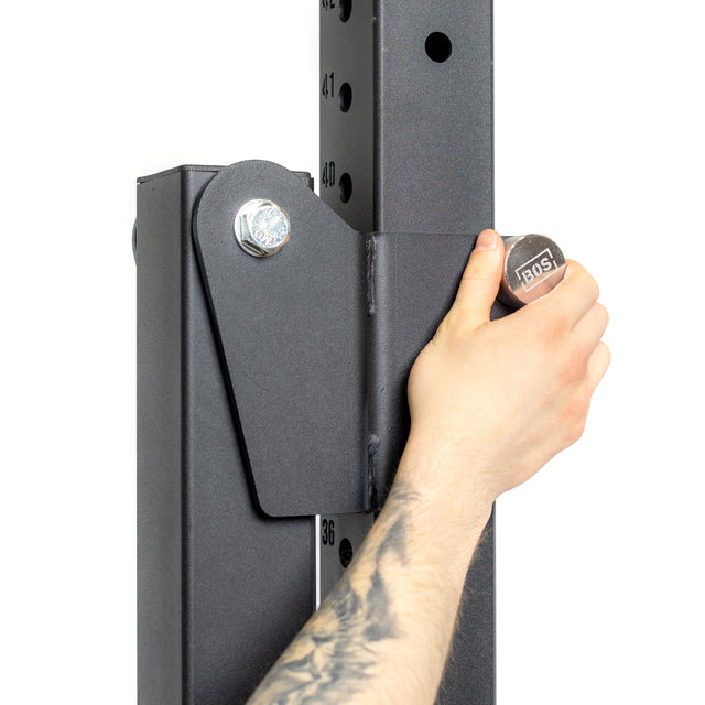 A person adjusts a Bells of Steel Magnetic Rack Attachment Pin on a black gym equipment rack. The anodized aluminum structure features numbered holes for height settings, and the person's tattooed forearm is visible.