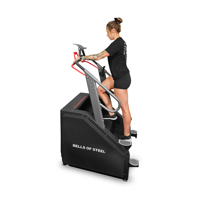 A person in athletic gear, with tattoos on their arms, uses a Bells of Steel Sisyphean Stepper. Dressed in a black t-shirt and shorts, they benefit from its handlebars and sturdy base that support various resistance levels for effective cardio workouts.