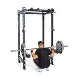 A man in athletic clothing squats beneath a barbell on his shoulders inside the Bells of Steel Manticore Four Post Power Rack Builder (3" x 3", 1" Holes). This sturdy home gym equipment is black and features modular components, offering various grip options and weight plates on each side of the barbell.