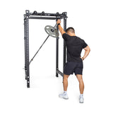 Dressed in a black sports outfit, an individual executes a landmine press exercise within the sophisticated Manticore Four Post Power Rack Builder (3" x 3", 1" Holes) from Bells of Steel. They skillfully grip a barbell with weight plates, making it an ideal setup for any home gym. With one arm extended and the other resting on their hip, the versatile modular components accentuate their workout routine.