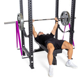 In a sleek home gym, a person is executing a bench press in the Manticore Four Post Power Rack Builder by Bells of Steel. Clad in a black outfit and white sneakers, they lift a barbell with precision. Resistance bands are skillfully attached to the modular components of the rack, enhancing their workout.