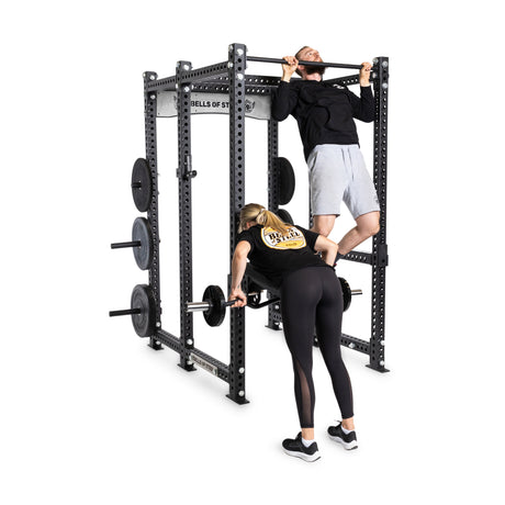 A man in black attire does a pull-up on the Bells of Steel Manticore Six Post Power Rack, while a woman in similar gear lifts a barbell from its modular components; weight plates are stacked nearby.