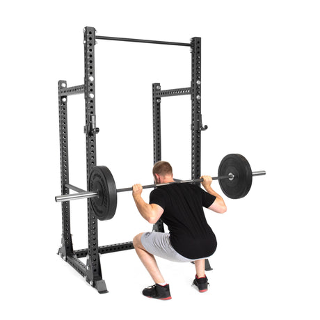 A man in a black shirt and gray shorts performs a squat with a barbell on his shoulders inside the Manticore Half Rack by Bells of Steel. This prebuilt power rack features modular components, supports up to 1,000 pounds, and ensures stability as he adds extra weight plates to the bar.