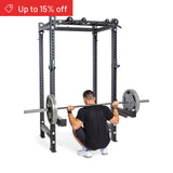 A man squats with a loaded barbell inside the Bells of Steel Manticore Four Post Power Rack Builder (3" x 3", 1" Holes). A red banner in the top left reads "Up to 15% off." The plain white background suits any home gym.