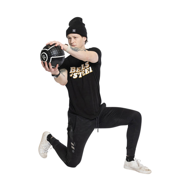 A person in athletic wear, including a black Bells of Steel shirt, black pants, white sneakers, and a beanie, lunges with a Mighty Grip Medicine Ball in hand. The medicine ball's superior grip enhances their fitness routine by adding stability to each movement.