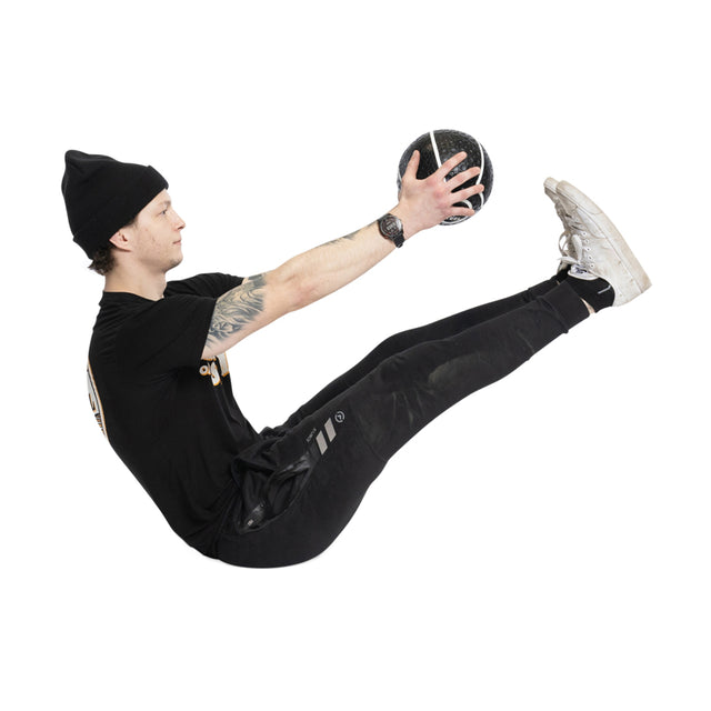 Wearing a black outfit and beanie, the person performs a V-sit with a Bells of Steel Mighty Grip Medicine Ball, renowned for its excellent grip. Sporting white sneakers and a visible arm tattoo, their fitness form stands out against the white background.